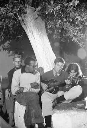 Leonard Cohen playing guitar on his right is Marianne Ihlen in Hydra, Greece where he bought a vacation home, October 1960. Photographer: James Burke Time Inc/Merlin.