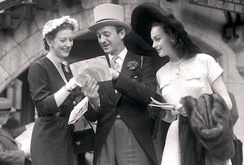 David Niven giving directions at the races with his wife, Hjordis on his right. Note his traditional charcoal striped trousers and voluptuous lapels.