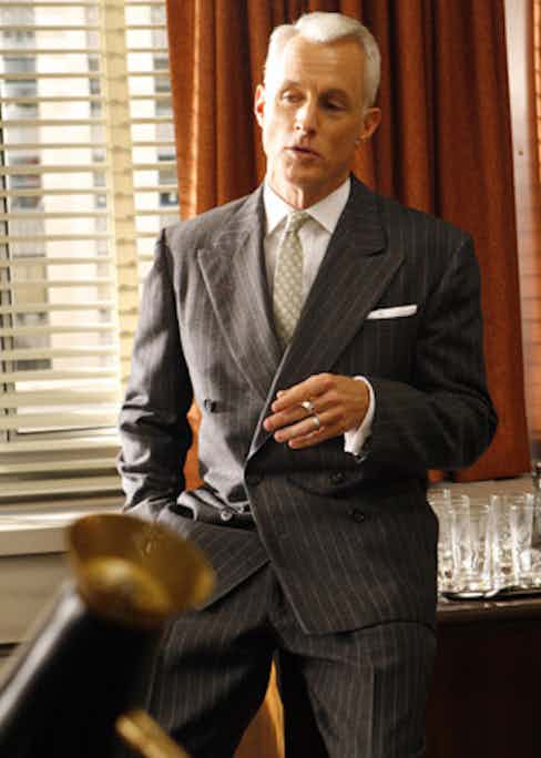 Roger Sterling makes a power play in a double-breasted charcoal suit with a wide pinstripe. The sloping shoulder and drape cut is true to the style of the time.