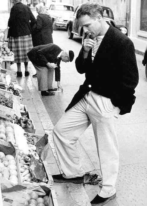 Buying fruit in the south of France, 1957. He wears wide-legged trousers, a boxy-cut blazer and a striped crew neck.