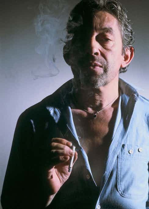 Opting for a casual denim shirt worn open and a silver necklace, Gainsbourg stands in his signature pose, cigarette in hand in Paris, 1985. Photograph by Jean-Jacques BERNIER/Gamma-Rapho via Getty Images.